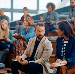 Eine Gruppe von Akademikern sitzt in einem modernen Hörsaal auf Holzbänken. Im Vordergrund unterhalten sich zwei Teilnehmende konzentriert, während andere Notizen in ihre Hefte schreiben.